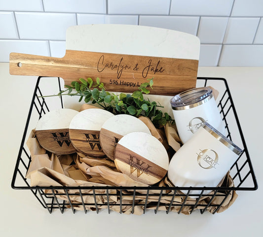 A custom gift basket containing a personalized marble acacia cutting board, round coasters, and wine tumblers, all engraved with names and dates, presented in a wire basket.